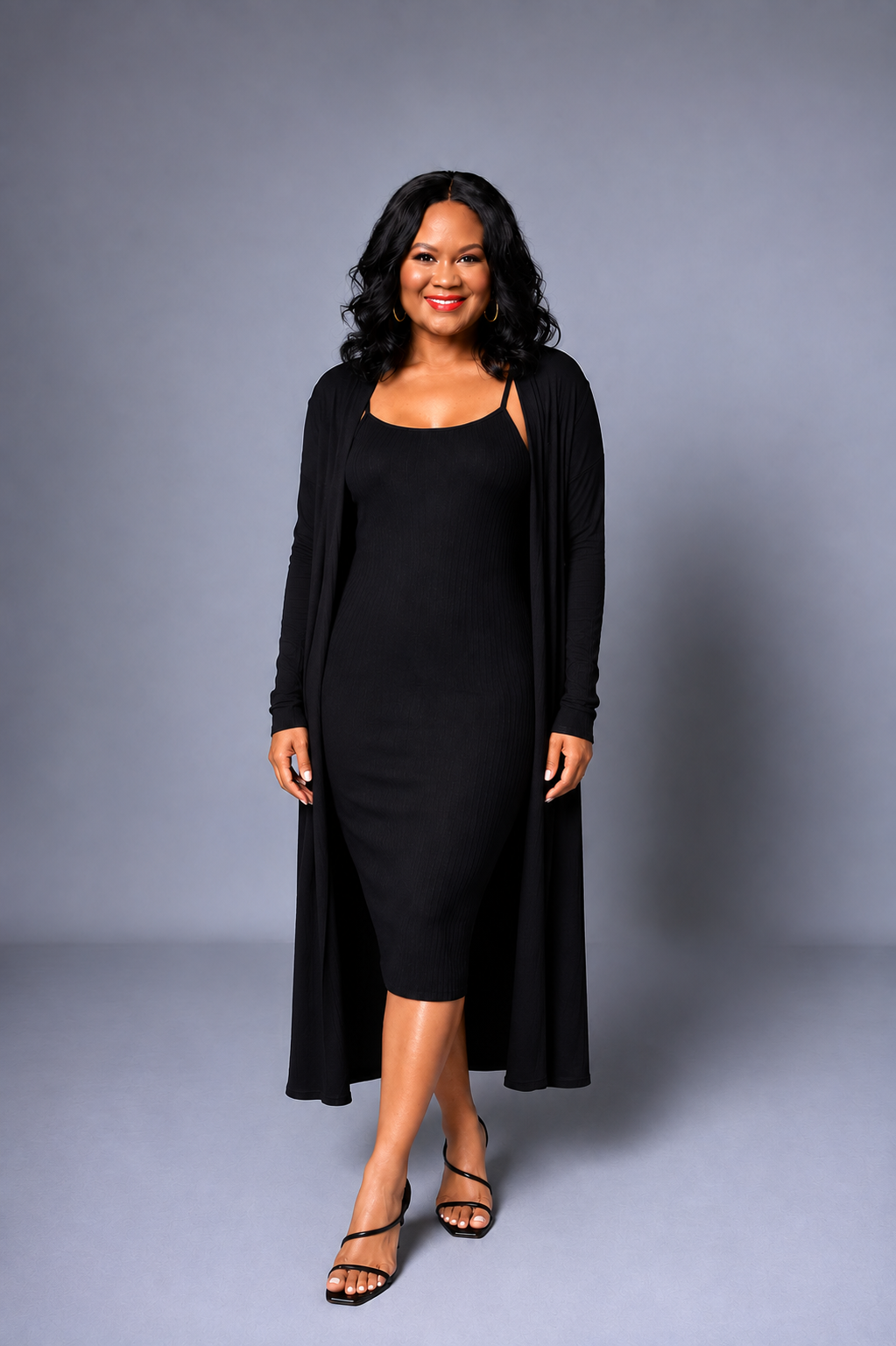 A woman modeling a black midi dress with a long open-front cardigan and black strappy heels, standing against a solid gray studio background.
