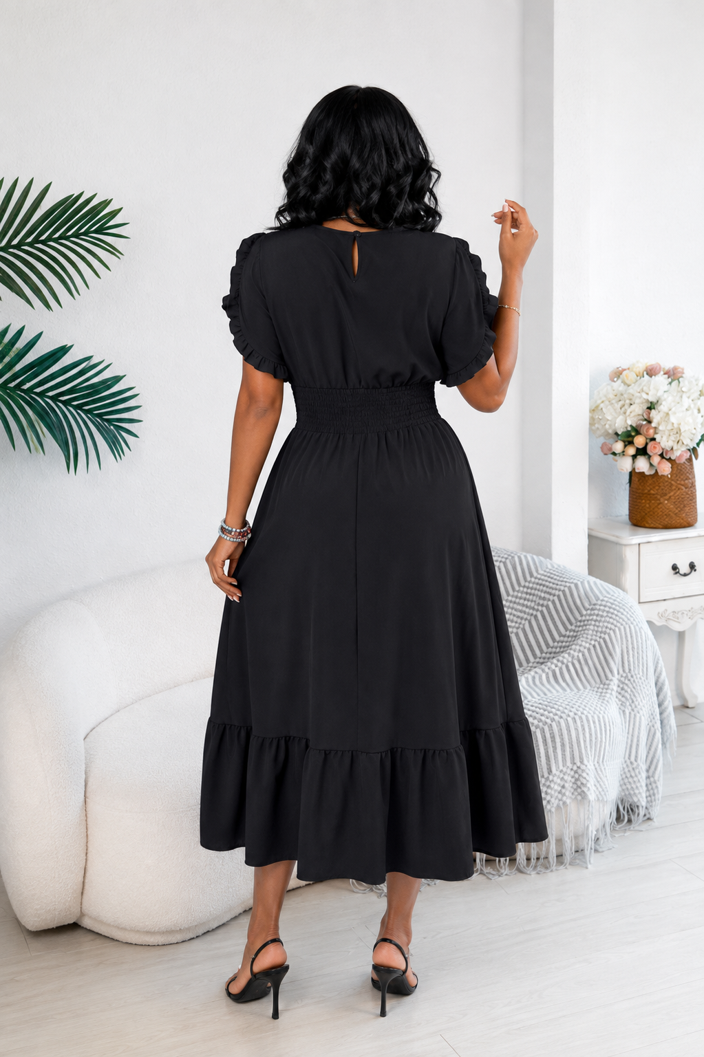 A woman wearing a black midi dress with puff sleeves and a tiered ruffle hem, shown from the back in a bright, modern room.