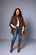 A woman modeling a brown open-front poncho-style coat over a white long-sleeve top, blue jeans, and brown knee-high boots, standing against a plain gray studio background.