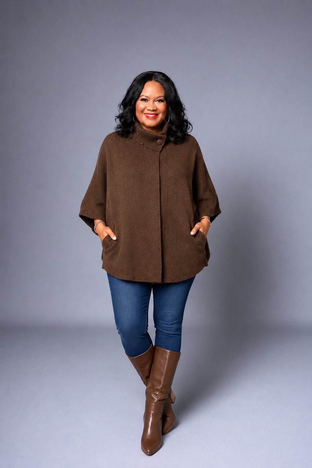 A woman modeling a brown poncho-style cardigan with front pockets, paired with blue jeans and brown knee-high boots, against a solid gray studio background.