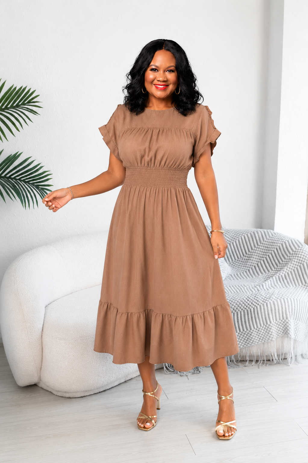 A woman modeling a brown midi dress with ruffled short sleeves, a smocked bodice, and a tiered skirt, standing in a bright, modern room with a white sofa and a striped throw blanket.