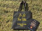 Black tote bag with yellow text on grass, featuring 'The Polished Wick Candle Company'.