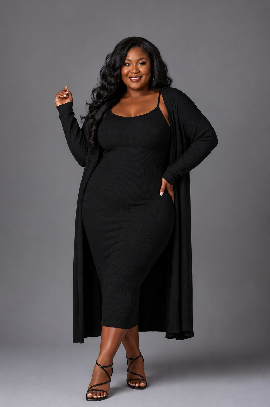 A woman modeling a black ribbed midi dress with a long-sleeve open-front cardigan draped over her shoulders, paired with black strappy heels, against a solid gray studio background.