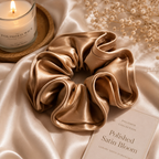 Gold satin scrunchie on a satin surface with a candle and flowers in the background