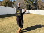 Person in graduation gown holding a black bag with text outdoors.