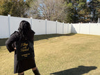 Person wearing a black coat and carrying a black bag with text in a backyard.