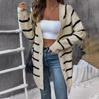 Woman wearing a beige cardigan with black stripes, white top, and blue jeans against a gray wall.