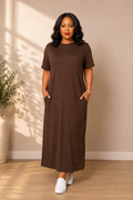 Woman wearing a brown dress standing in a room with a plant and wooden floor.