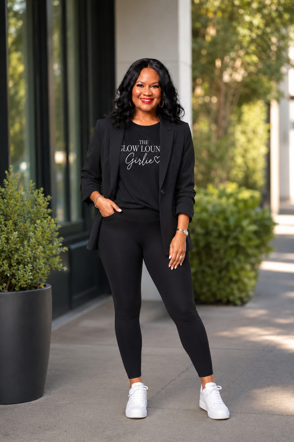 A woman stands outdoors wearing a black outfit consisting of a graphic T-shirt that reads 'The Slow Loun Garber', a black blazer, black leggings, and white sneakers.
