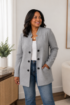 A woman wearing a light gray double-breasted blazer with black buttons, a white top, and blue jeans, standing in a bright, modern room.