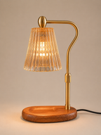 A modern table lamp with a curved brass metal arm, a ribbed clear glass shade, and a round wooden base, shown lit against a neutral beige background.