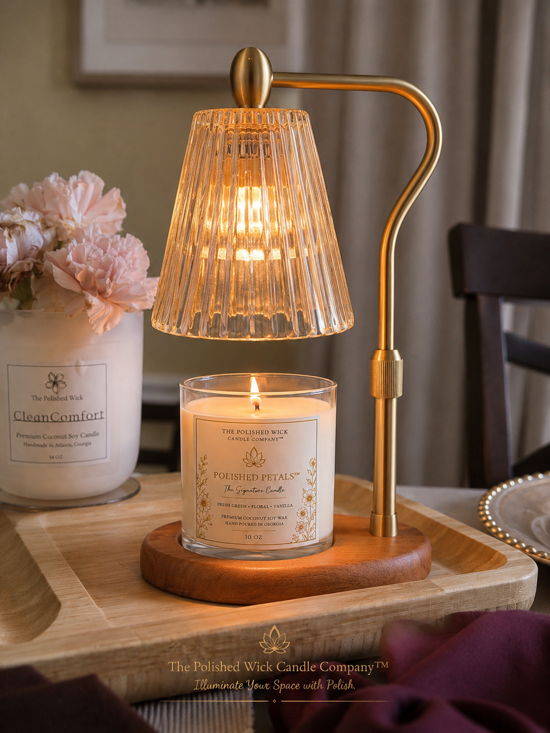 A lit candle in a clear ribbed glass jar sits on a wooden base next to a brass table lamp with a pleated glass shade, styled on a wooden tray with a vase of pink flowers in a home setting.