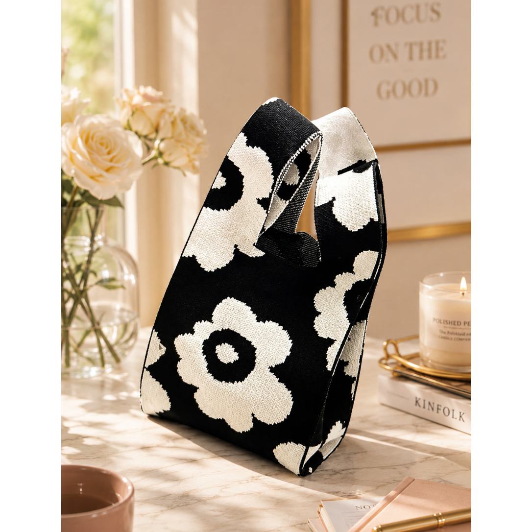 A black and white knit tote bag with a bold floral pattern, styled on a marble tabletop next to a vase of roses, a lit candle, and a notebook.