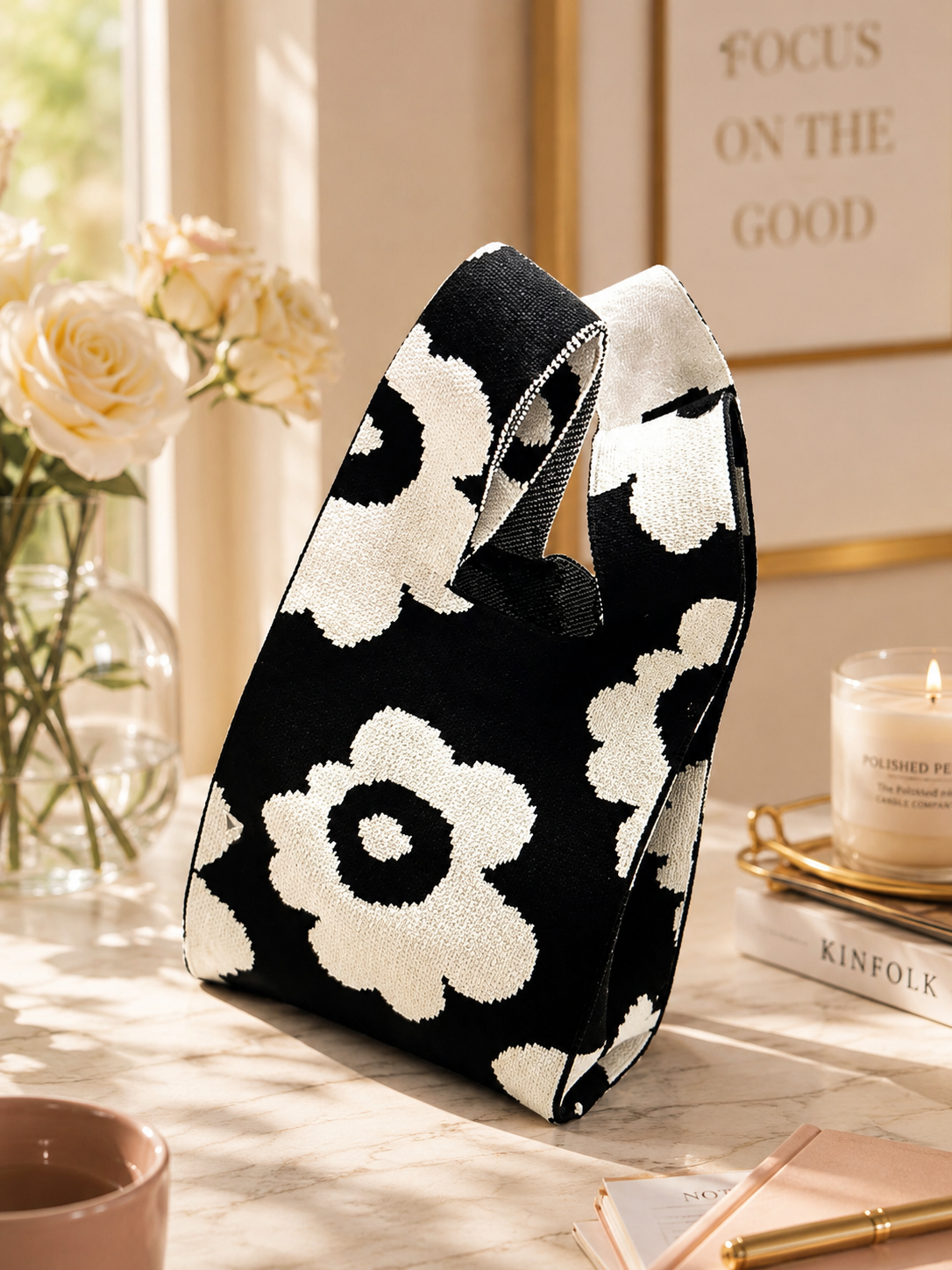 A black and white knit tote bag with a bold floral pattern, styled on a marble tabletop next to a vase of roses, a lit candle, and a notebook.