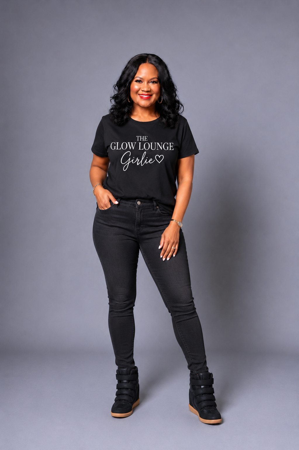 A woman modeling a black graphic t-shirt with 'THE GLOW LOUNGE Girlie' printed in white script, paired with black skinny jeans and black wedge boots, against a solid gray studio background.