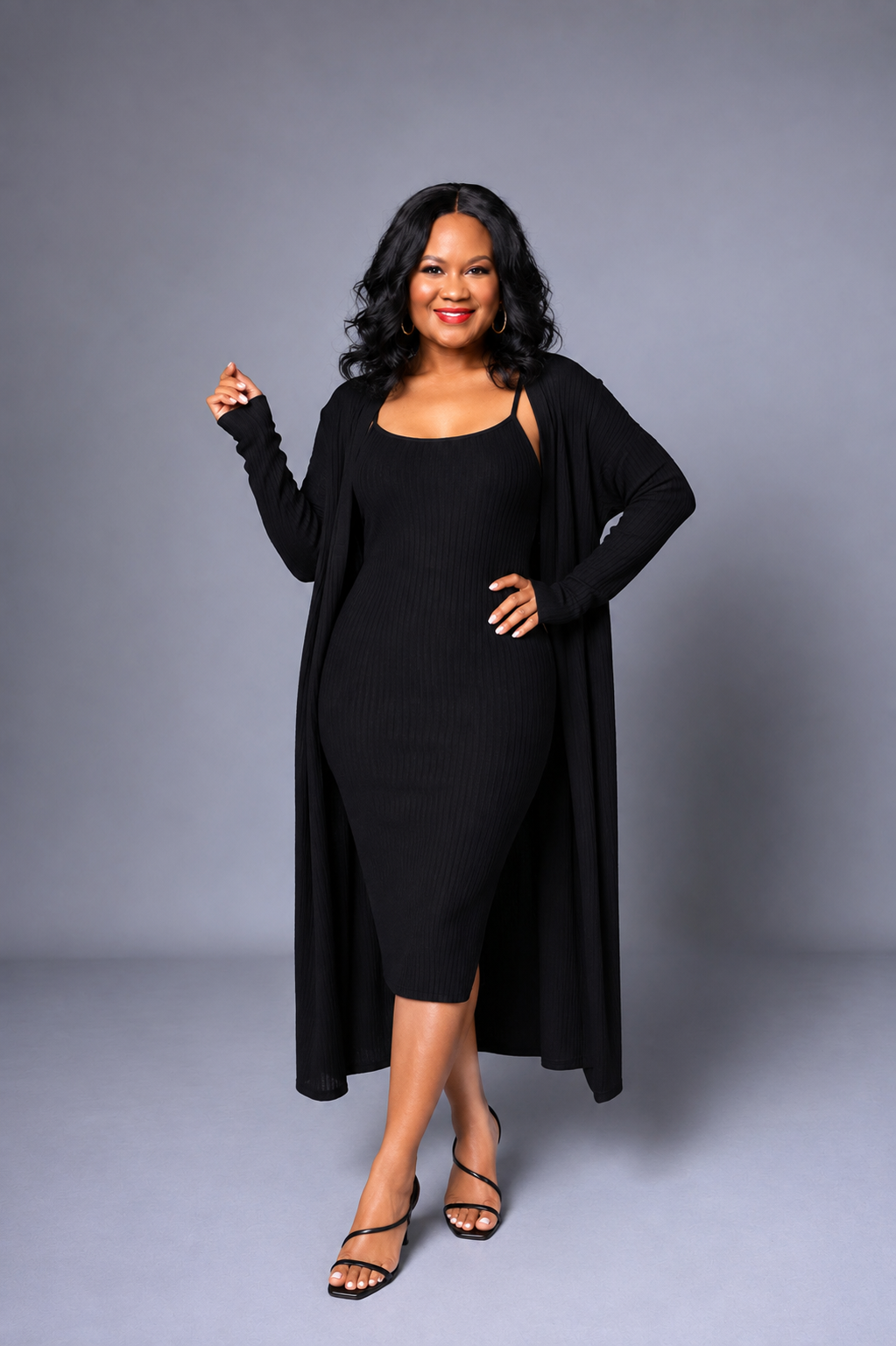 A woman modeling a black ribbed midi dress with a long open-front cardigan, styled with black strappy heels against a solid gray studio background.